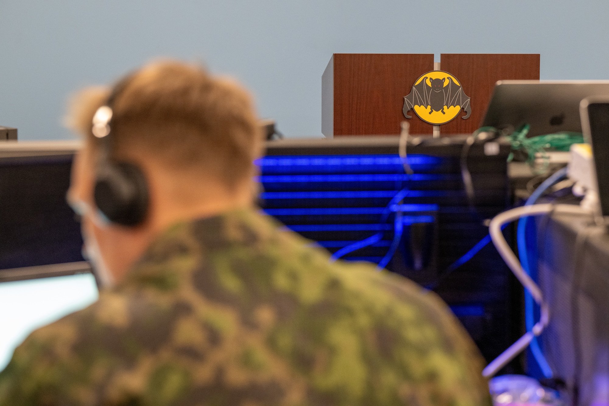 Finnish Air Force member in the foreground, out of focus, with headphones on looking at computer, 185th COS bat logo in background in focus