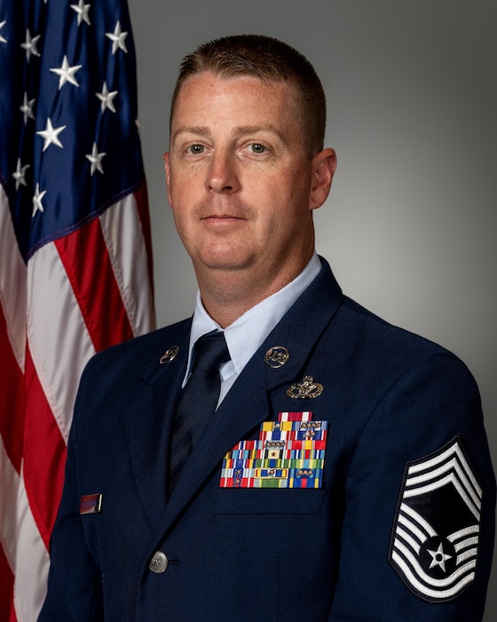 A man in military uniform stands in front of an American Flag
