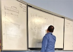 A man in a blue collard shirt writes mathematical equations on a whiteboard.