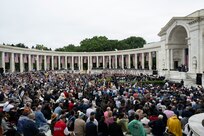Thousands of people are gathered in a large circular amphitheater which has white columns and large stage. There are U.S. flags hanging between each of the columns.