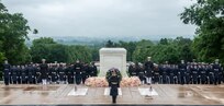 Dozens of military members from various branches of the armed forces are standing in formations wearing raincoats and hats near the Tomb of the Unknown Soldier. There is a large green wreath and multicolored flowers around the Tomb.