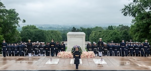 Dozens of military members from various branches of the armed forces are standing in formations wearing raincoats and hats near the Tomb of the Unknown Soldier. There is a large green wreath and multicolored flowers around the Tomb.