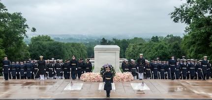 Dozens of military members from various branches of the armed forces are standing in formations wearing raincoats and hats near the Tomb of the Unknown Soldier. There is a large green wreath and multicolored flowers around the Tomb.