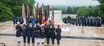 Military members are in various formations near the Tomb of the Unknown Soldier. In front of and facing the tomb is a group of nine military members from each other branches carrying the U.S. flag and the flags for each of the six branches of the armed forces.