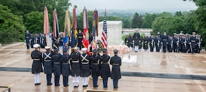 Military members are in various formations near the Tomb of the Unknown Soldier. In front of and facing the tomb is a group of nine military members from each other branches carrying the U.S. flag and the flags for each of the six branches of the armed forces.