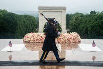 An Army soldier dressed in dark blue ceremonial uniform with raincoat, cap and white gloves is marching on a black mat near the white marble-clad Tomb of the Unknown Soldier. In front of the tomb in the background are small U.S. Flag and a large arrangement of multicolored flowers at the base of the Tomb.