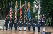 Military members wearing ceremonial uniforms with raincoats and caps are presenting the colors, which includes the U.S. Flag and the flag for each of the U.S. Armed Forces, during an outdoor ceremony at the Tomb of the Unknown Soldier.