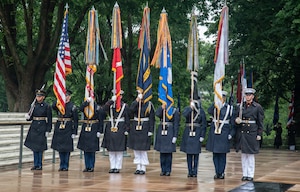 Military members wearing ceremonial uniforms with raincoats and caps are presenting the colors, which includes the U.S. Flag and the flag for each of the U.S. Armed Forces, during an outdoor ceremony at the Tomb of the Unknown Soldier.