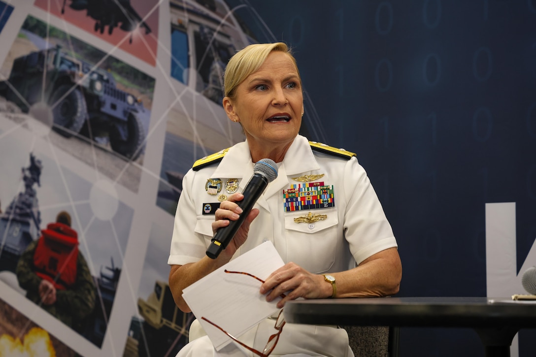 A woman speaks into a microphone. She is wearing a dress white Navy uniform and is seated on a small stage with her arm resting on a table.