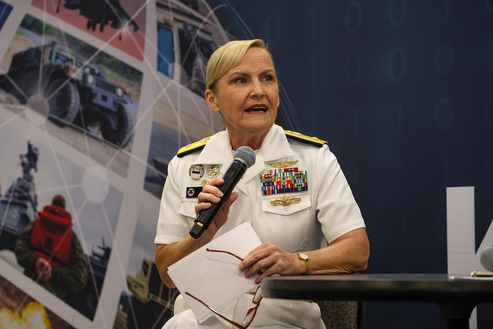 A woman speaks into a microphone. She is wearing a dress white Navy uniform and is seated on a small stage with her arm resting on a table.