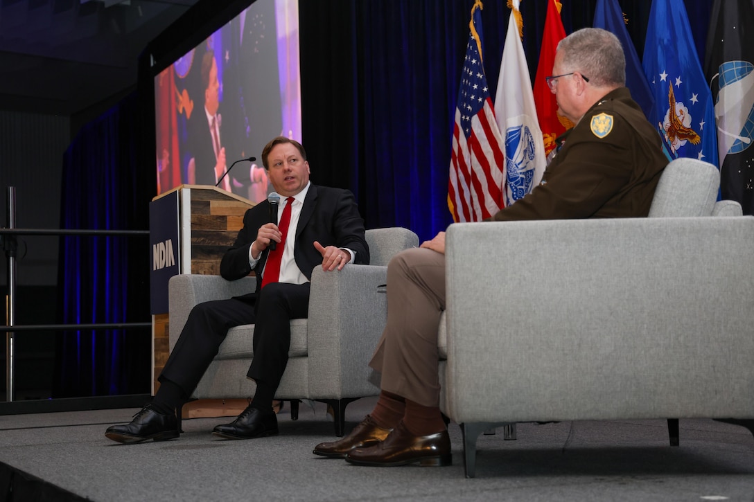 A man in a dark business suit sits in a chair on a ballroom stage. He is holding a microphone and is chatting with a man in a Army dress uniform also seated on stage.