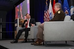 A man in a dark business suit sits in a chair on a ballroom stage. He is holding a microphone and is chatting with a man in a Army dress uniform also seated on stage.