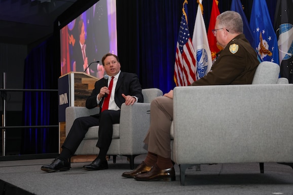 A man in a dark business suit sits in a chair on a ballroom stage. He is holding a microphone and is chatting with a man in a Army dress uniform also seated on stage.
