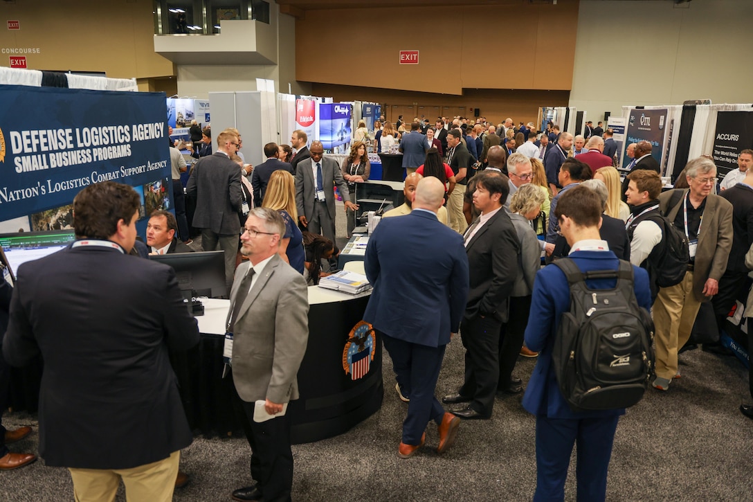 A large group of people chat in an aisleway on a exhibit hall floor at several booths with the DLA Office of Small Business Programs booth displayed prominently in the foreground.