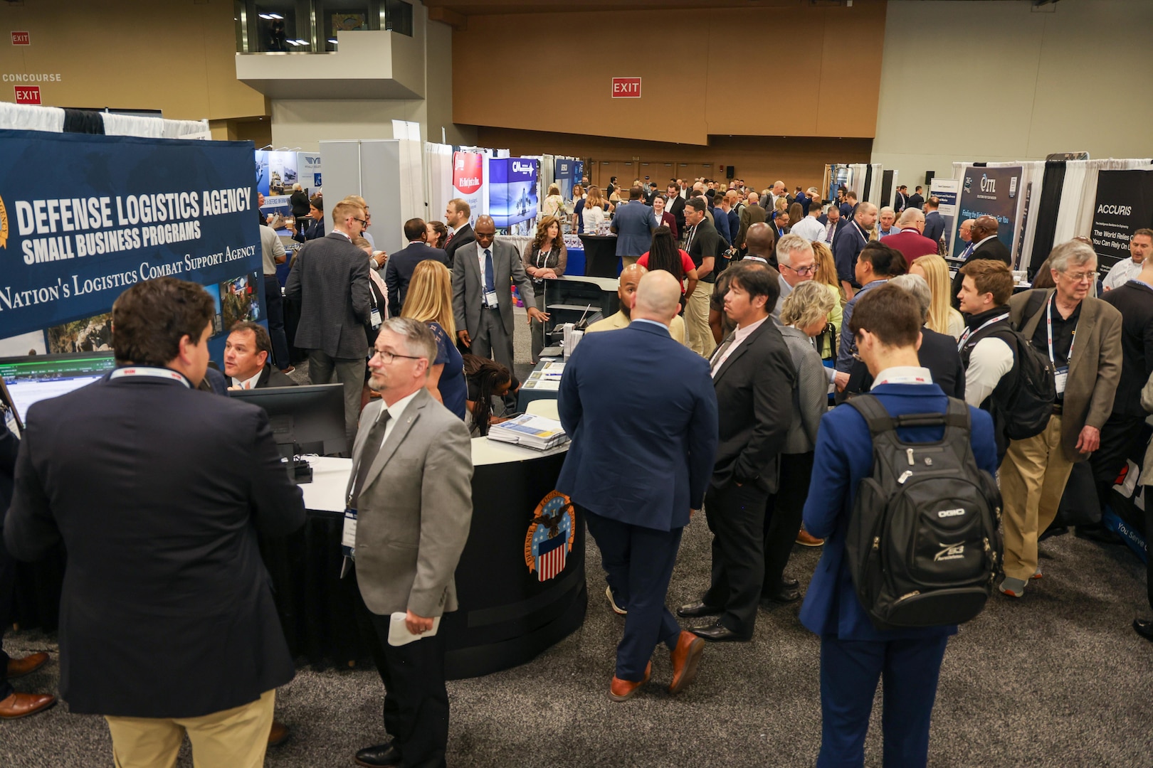 A large group of people chat in an aisleway on a exhibit hall floor at several booths with the DLA Office of Small Business Programs booth displayed prominently in the foreground.