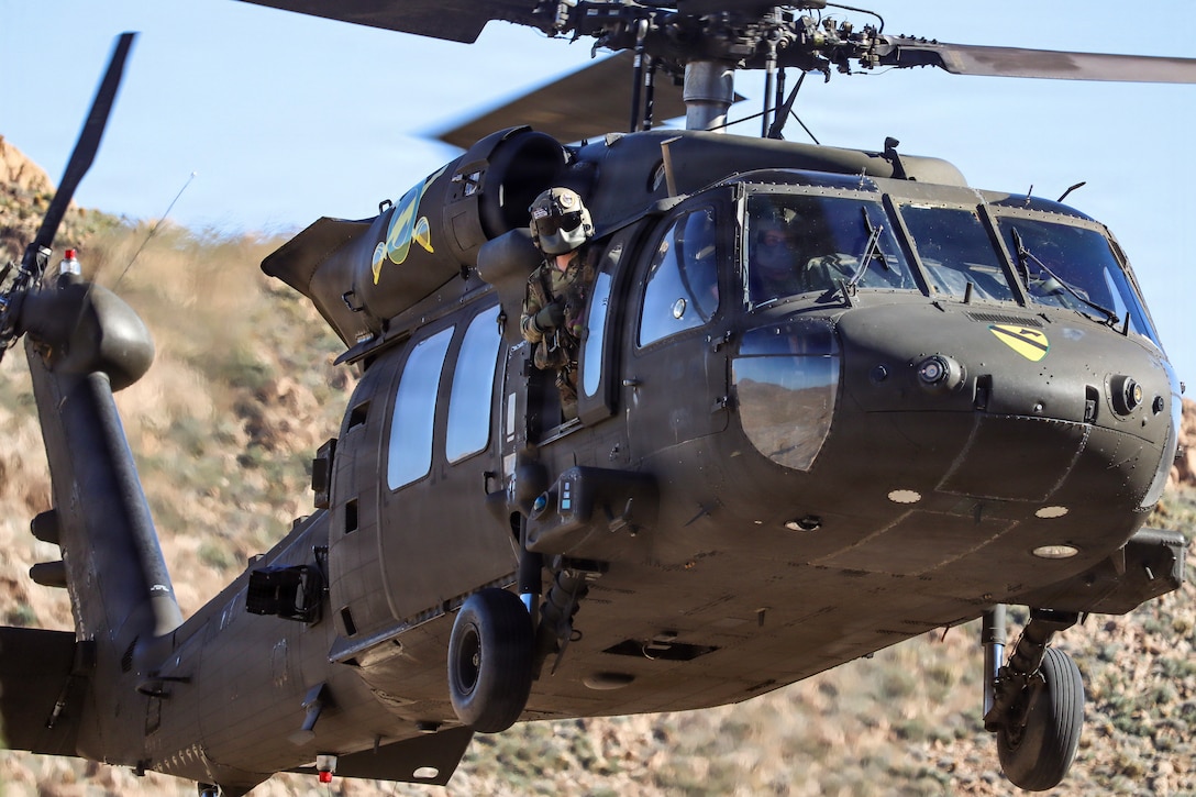 A soldier in a flight mask looks out from the side of a helicopter flying over arid terrain.
