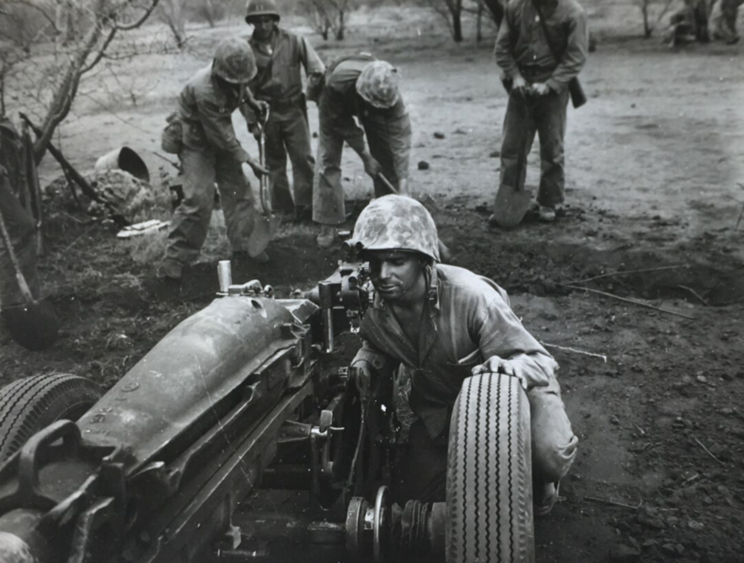 A black and white image of a Marine squatting down behind a large piece of equipment on wheels looking into a scope.