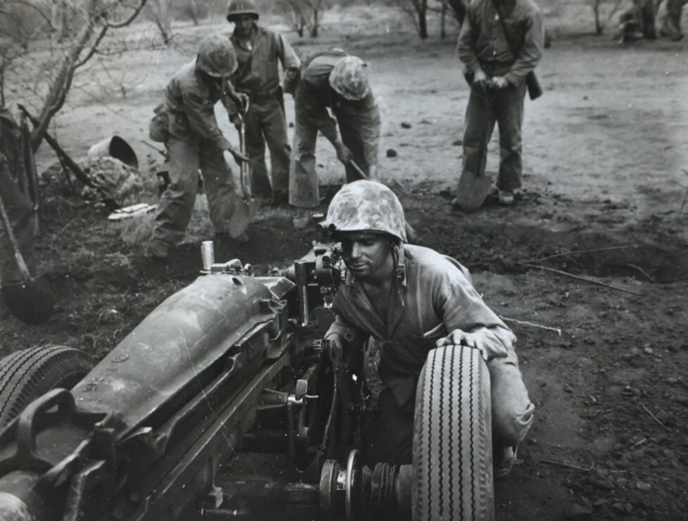 A black and white image of a Marine squatting down behind a large piece of equipment on wheels looking into a scope.