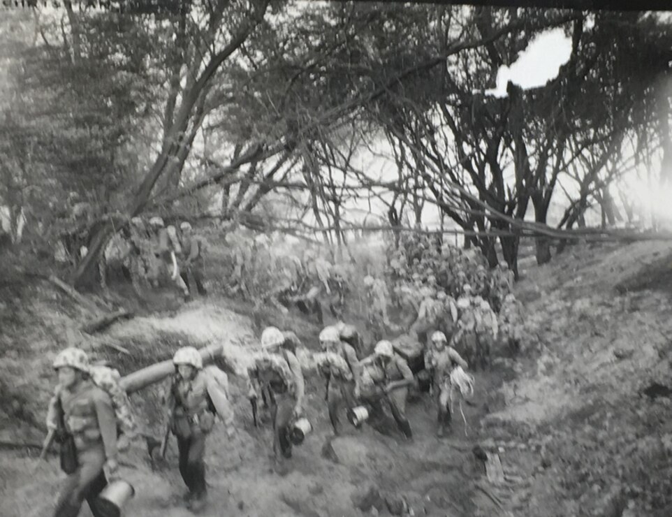 A black and white image showing a battalion of Marines carrying equipment walking through a path in a forest, inland away from a bay.