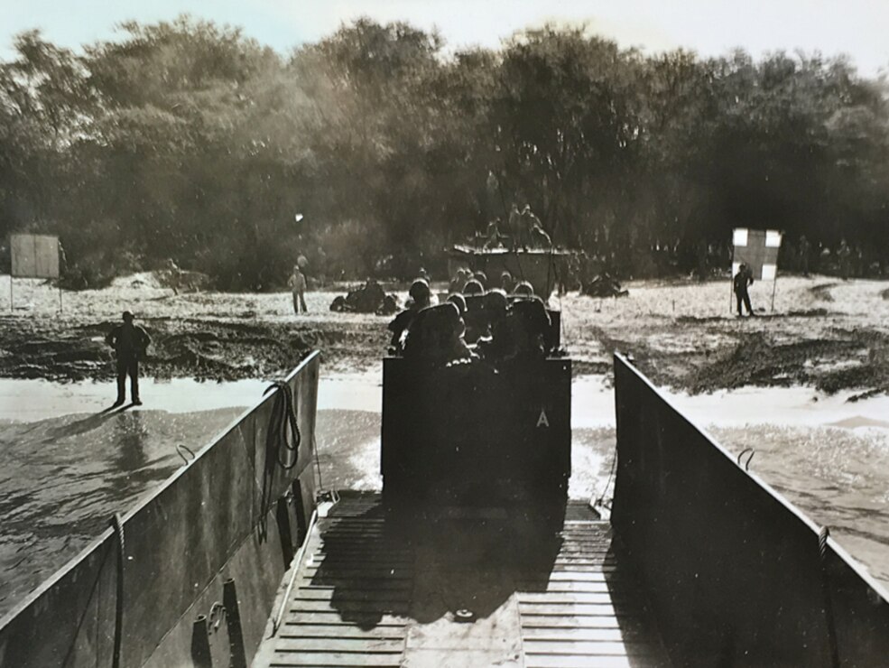 Black and white image with high contrast. Amphibious WWII vehicle exits the water and on to the beach. A thick row of trees in background.