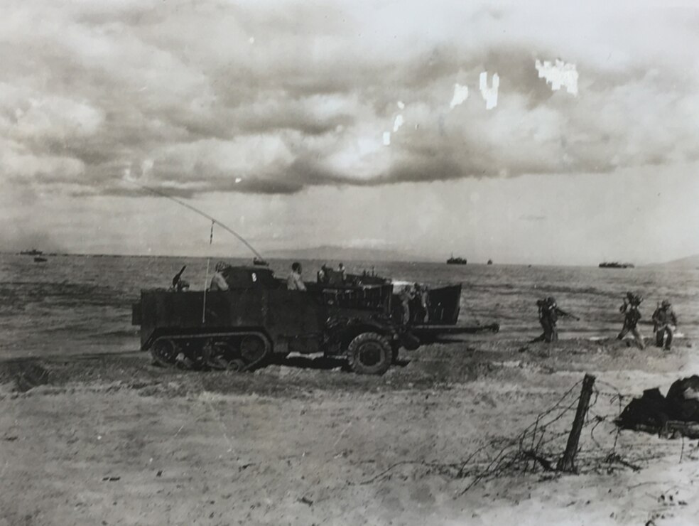 Black and white image of one WWII amphibious vehicle on land with three military members following on foot. Low clouds hang in the distance.