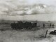 Black and white image of one WWII amphibious vehicle on land with three military members following on foot. Low clouds hang in the distance.