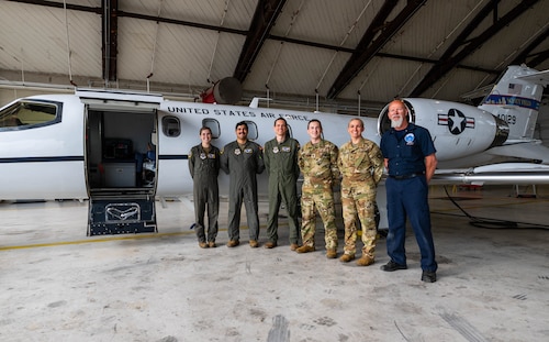 A group of Airmen, and a civilian on the far right, stand with their hands behind their back, in front of a C-21 jet.