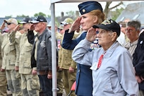 A veteran in a black hat salutes among a standing audience during a ceremony.