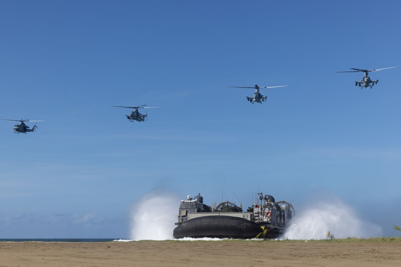 Four military aircraft fly over a military watercraft as it lands on a beach.