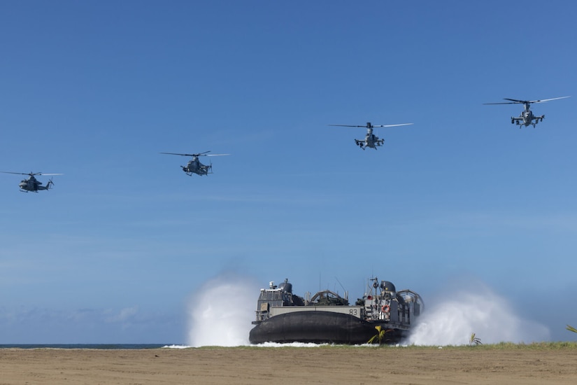 Four military aircraft fly over a military watercraft as it lands on a beach.