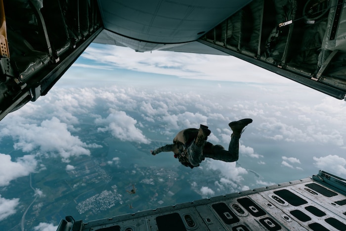 A person wearing a camouflage military uniform and parachute jumps out the back of a military aircraft into a cloudy sky.