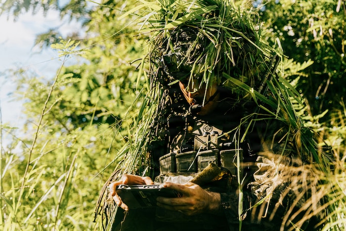 A man wearing a camouflage military uniform looks at a screen while covered in camouflage debris in a jungle.
