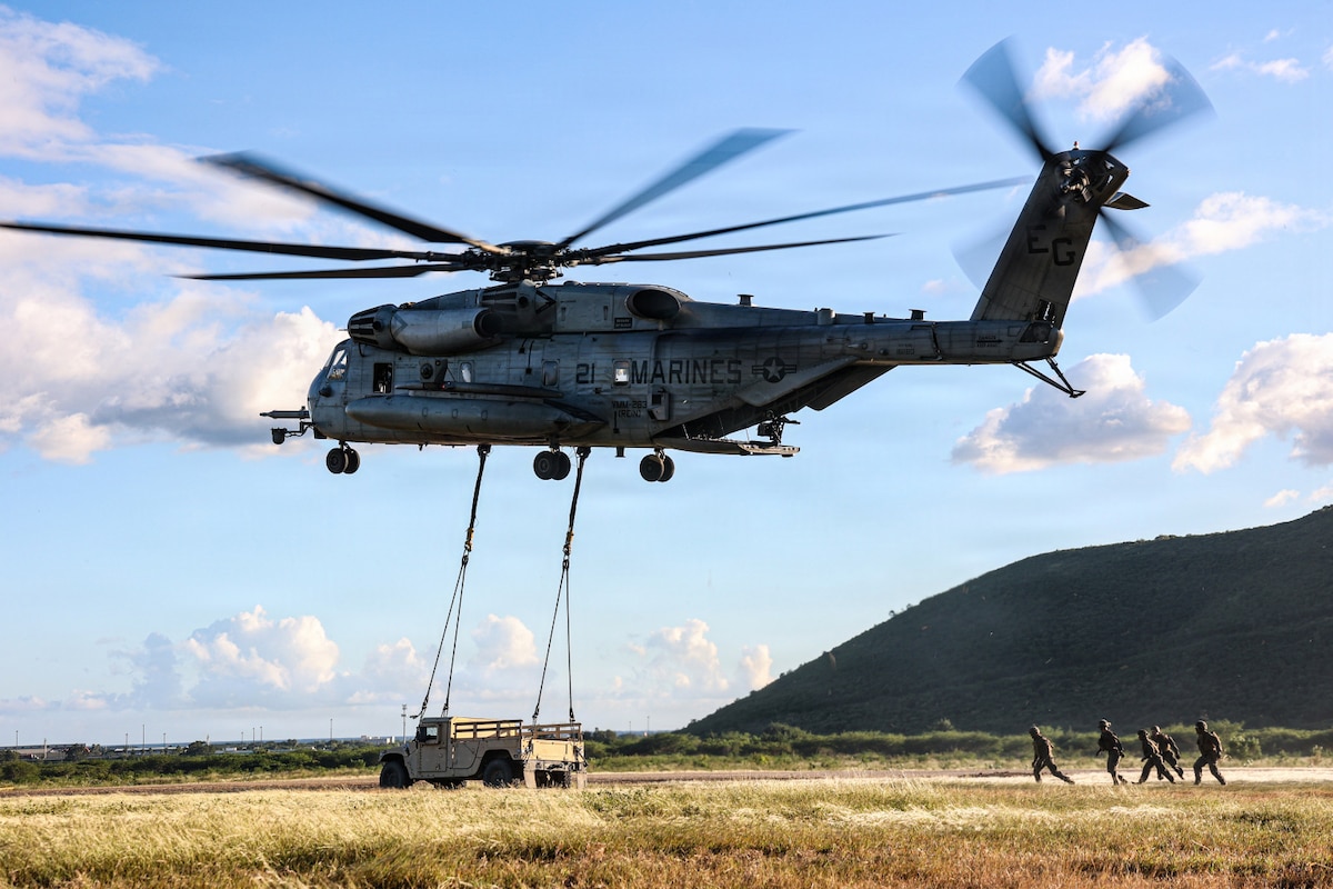 A military helicopter hovers above the ground while preparing to lift a Humvee; there are five people wearing camouflage military uniforms observing on the ground.