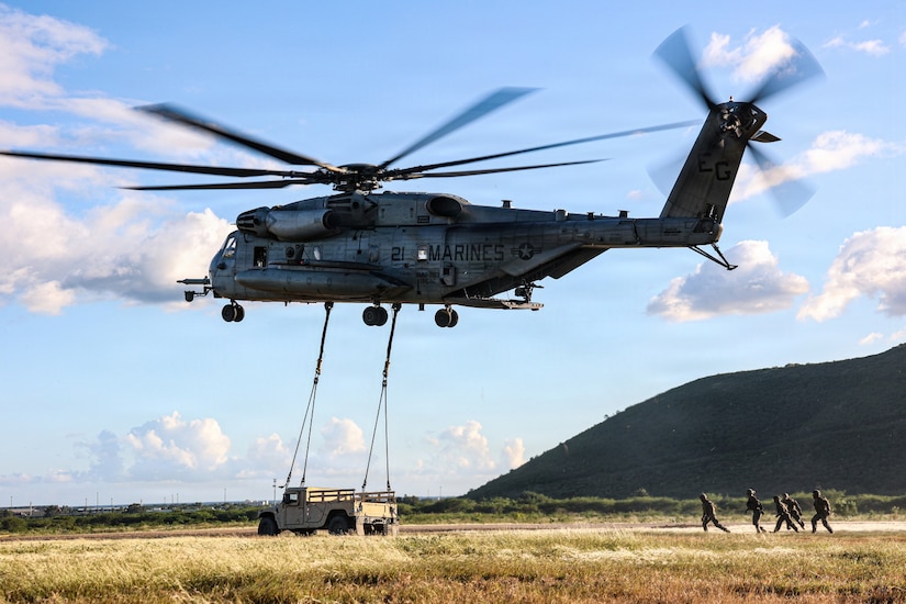 A military helicopter hovers above the ground while preparing to lift a Humvee; there are five people wearing camouflage military uniforms observing on the ground.
