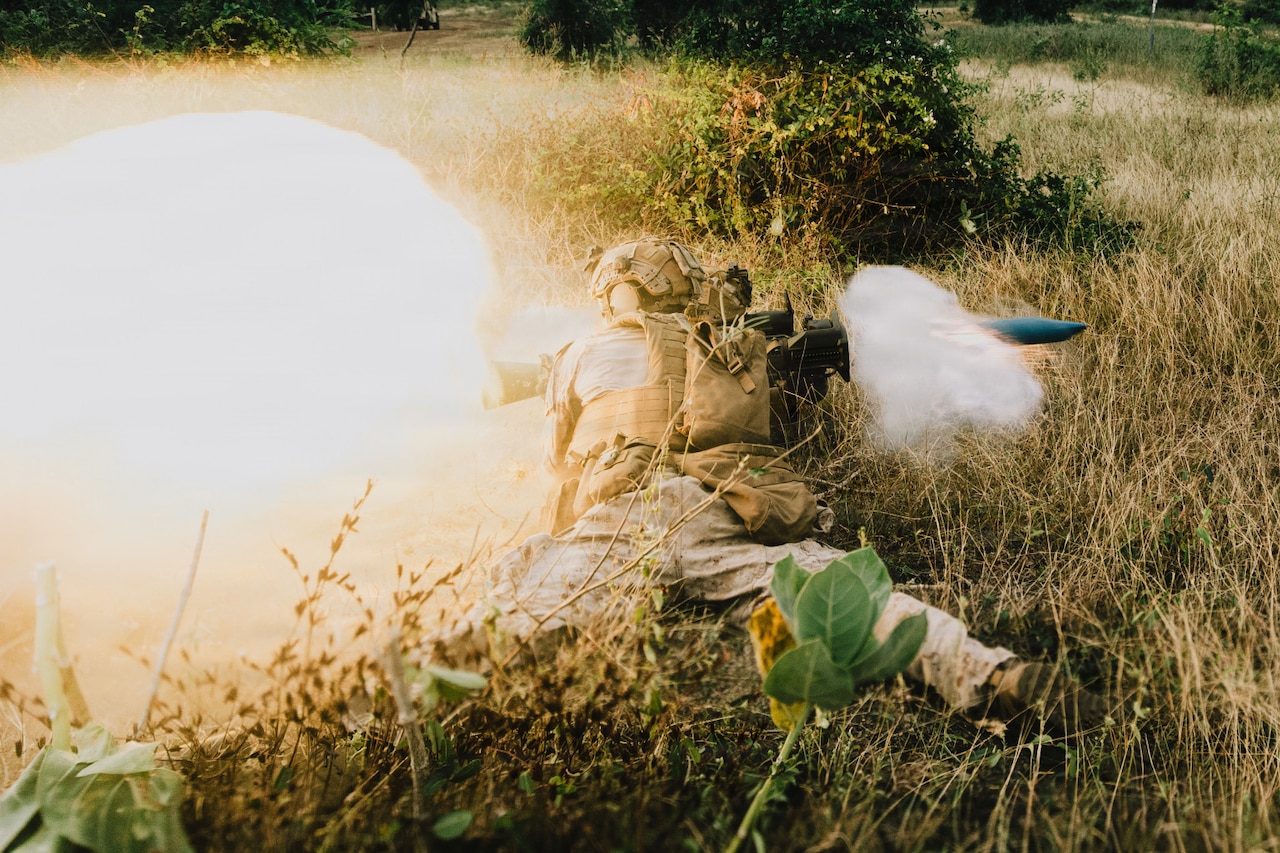 A person wearing tactical gear and a camouflage military uniform lies on the ground in thick brush while firing a rocket.