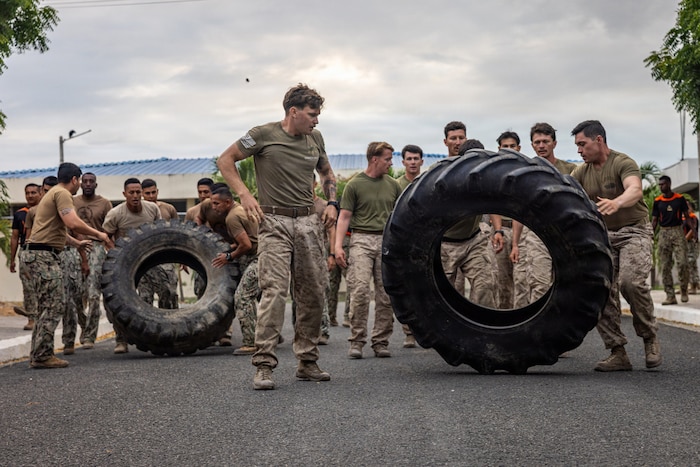Two groups of men wearing camouflage military uniforms flip large tires over on a road.