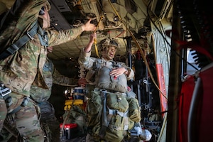 A man in a camouflage military uniform and jump gear prepares to jump out of a military aircraft. Another man in a camouflage military uniform reaches up to grab a jump line.
