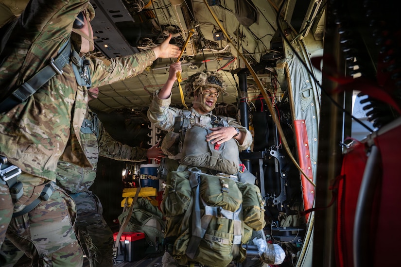A man in a camouflage military uniform and jump gear prepares to jump out of a military aircraft. Another man in a camouflage military uniform reaches up to grab a jump line.