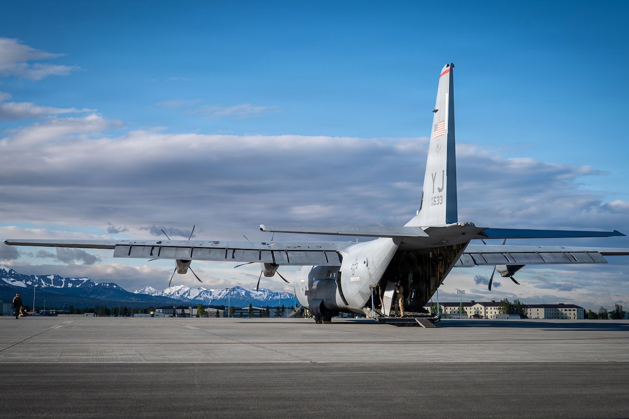 A military cargo airplane sits on a flight line. Buildings and mountains are in the background.