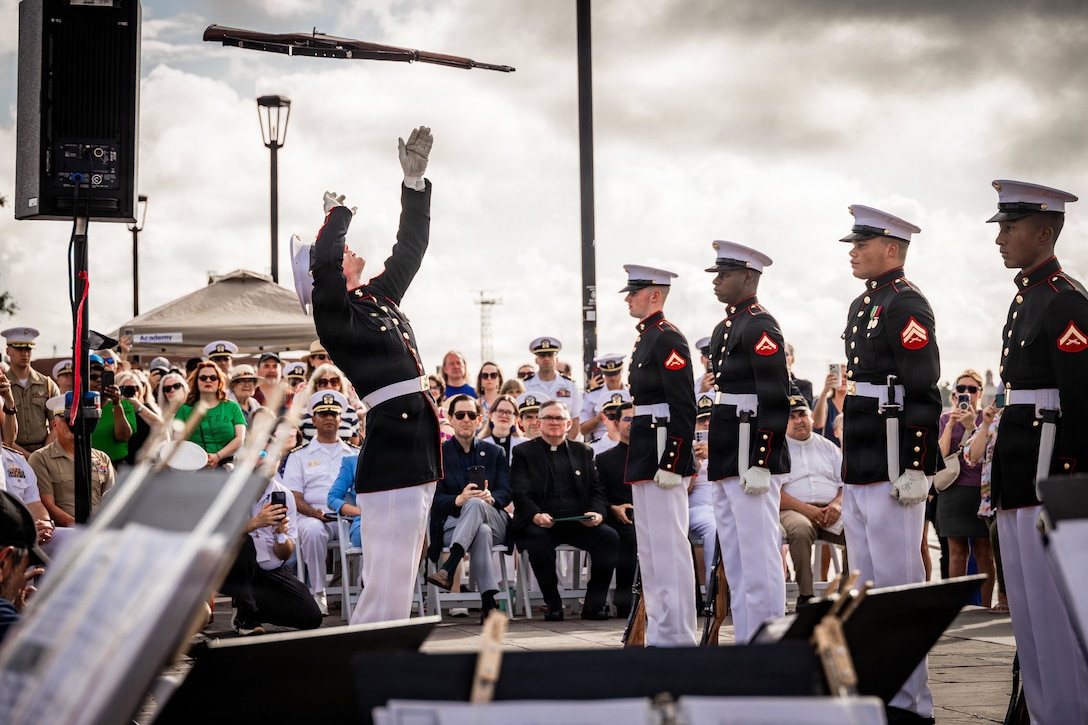 A Marine in ceremonial dress tosses a rifle as fellow Marines stand at attention during a ceremony while an audience watches under a cloudy sky.