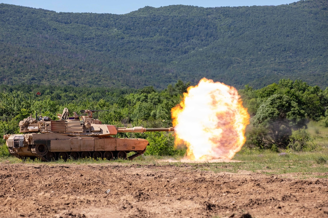 Soldiers fire a tank in a field, creating a large, orange fireball with a mountain in the background.