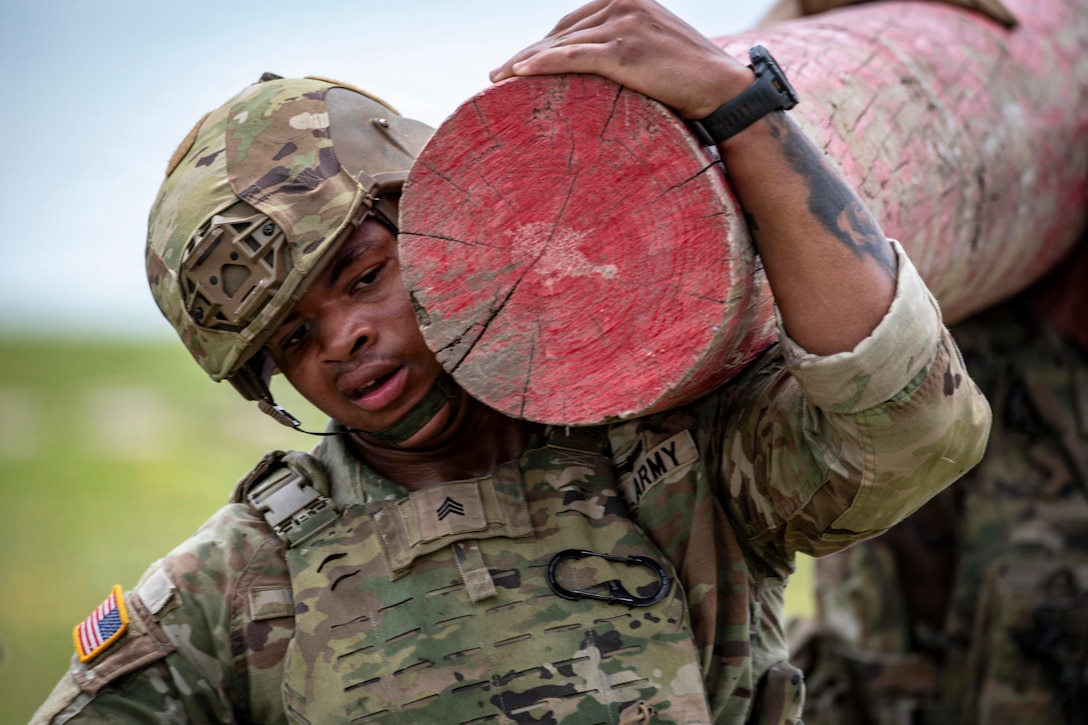A close-up of a soldier wearing a helmet carrying a log on their shoulder.