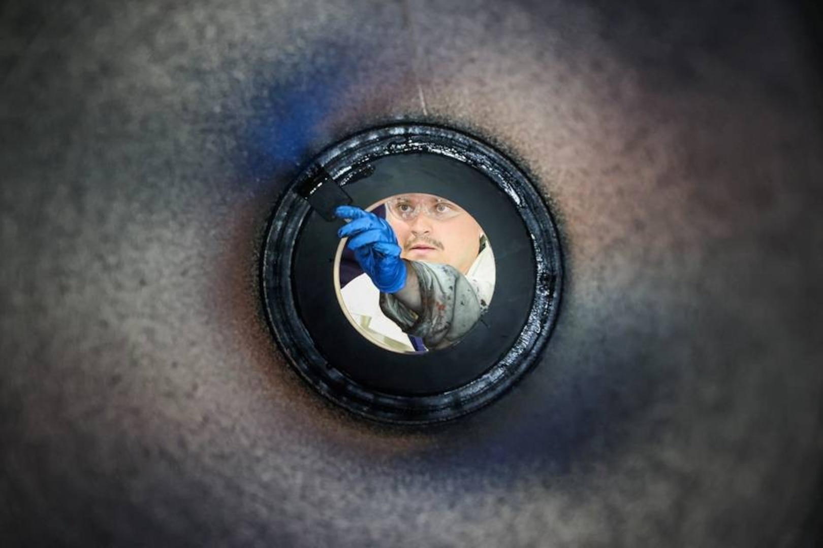 Photograph taken from inside a cylindrical pipe looking outward, showing a person wearing blue gloves and holding a tool, performing completion of lining of the casing. The circular frame of the pipe creates a tunnel effect, focusing attention on the worker and the task being performed.