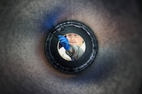 Photograph taken from inside a cylindrical pipe looking outward, showing a person wearing blue gloves and holding a tool, performing completion of lining of the casing. The circular frame of the pipe creates a tunnel effect, focusing attention on the worker and the task being performed.
