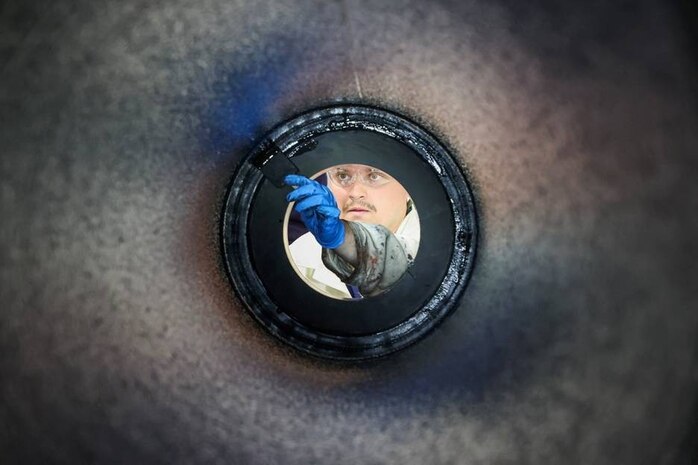 Photograph taken from inside a cylindrical pipe looking outward, showing a person wearing blue gloves and holding a tool, performing completion of lining of the casing. The circular frame of the pipe creates a tunnel effect, focusing attention on the worker and the task being performed.