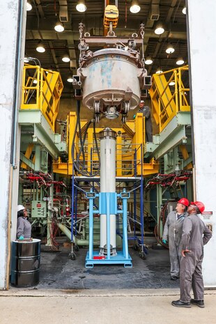 Photograph of an industrial facility showing a large cylindrical metal motor suspended by a crane above a vertical support structure, surrounded by yellow platforms and machinery. Three workers in gray uniforms and red helmets observe the setup.