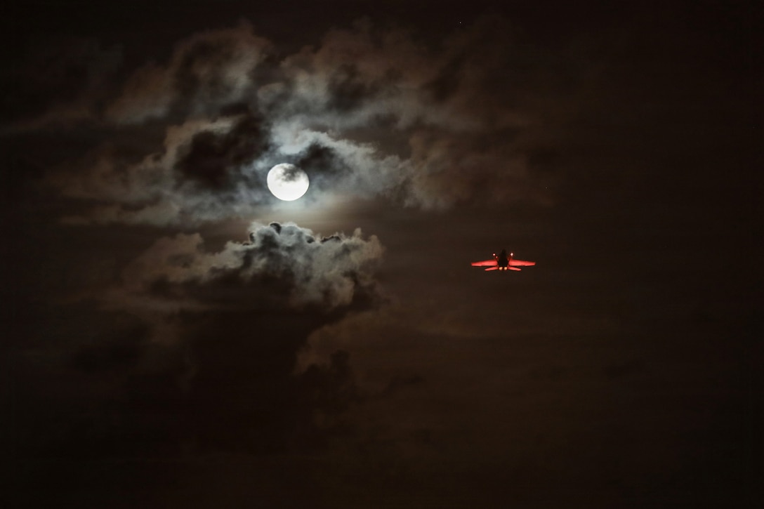 An aircraft illuminated by red light flies in a dark sky near a partially cloud-covered moon.