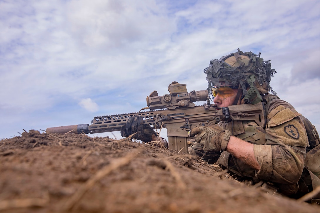 A soldier in tactical gear aims a rifle on a pile of dirt against a blue sky.