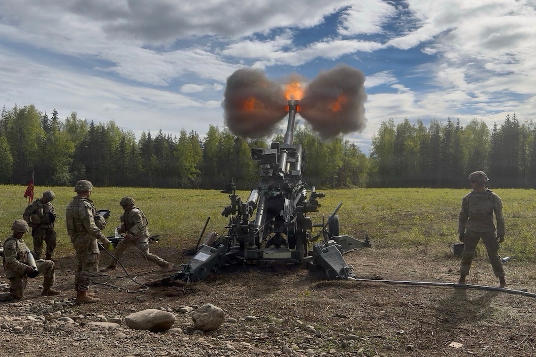 Soldiers in tactical gear fire a weapon in a field, creating a smoky fireball against a blue sky.