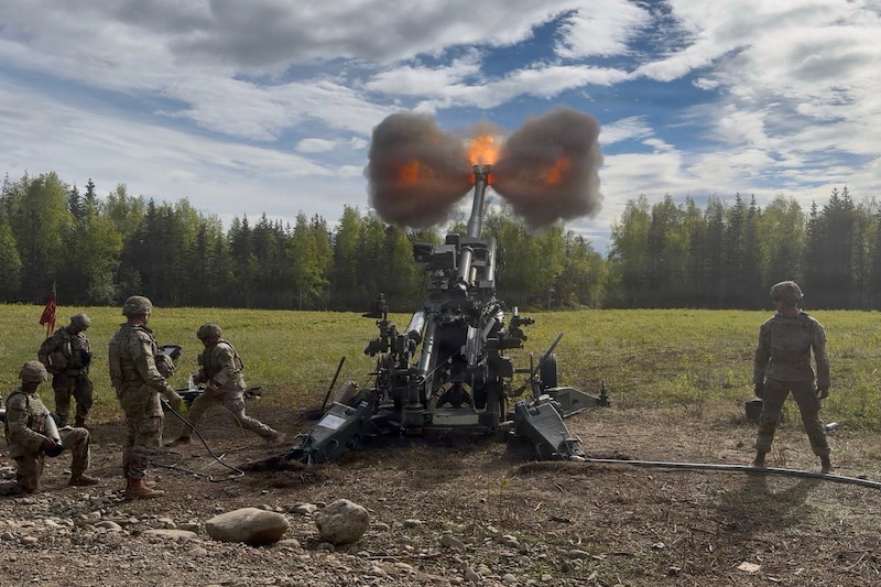 Six soldiers work in a grass field by a howiter they are firing, creating tufts of smoke and flames.
